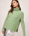 Woman wearing a sage Claudia turtleneck sweater with wide rib detail and side slits.