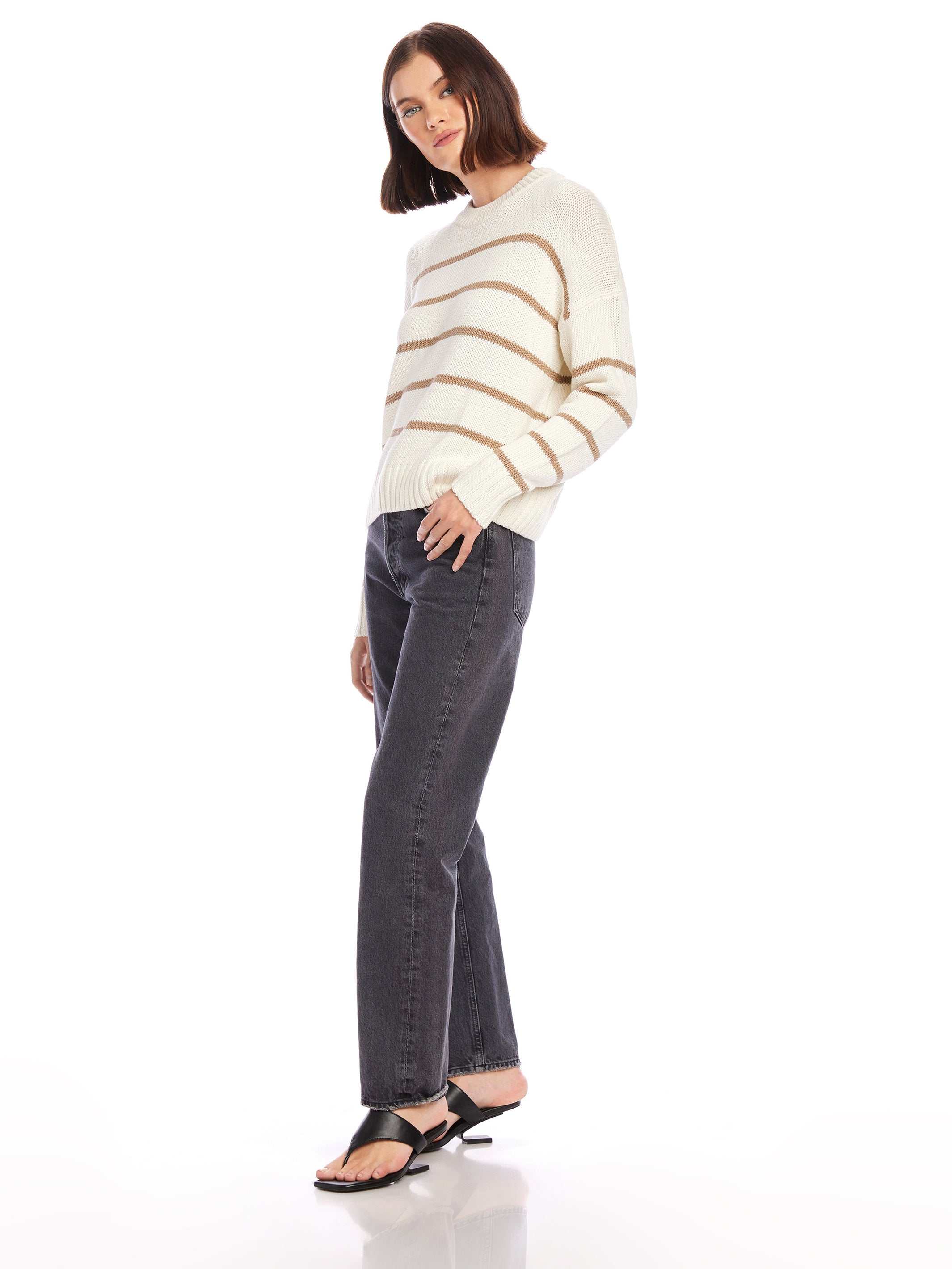 Woman wearing Luca Crewneck Sweater with beige stripes, paired with black jeans and sandals, posing against white background
