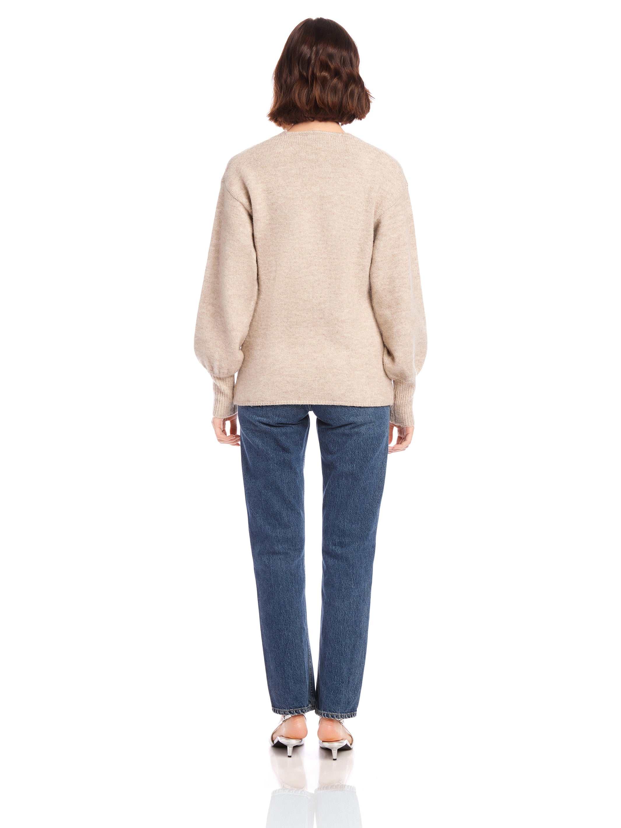 Back view of a woman wearing a beige Lucy Cardigan with long blouson sleeves and wide cuffs paired with blue jeans and heels