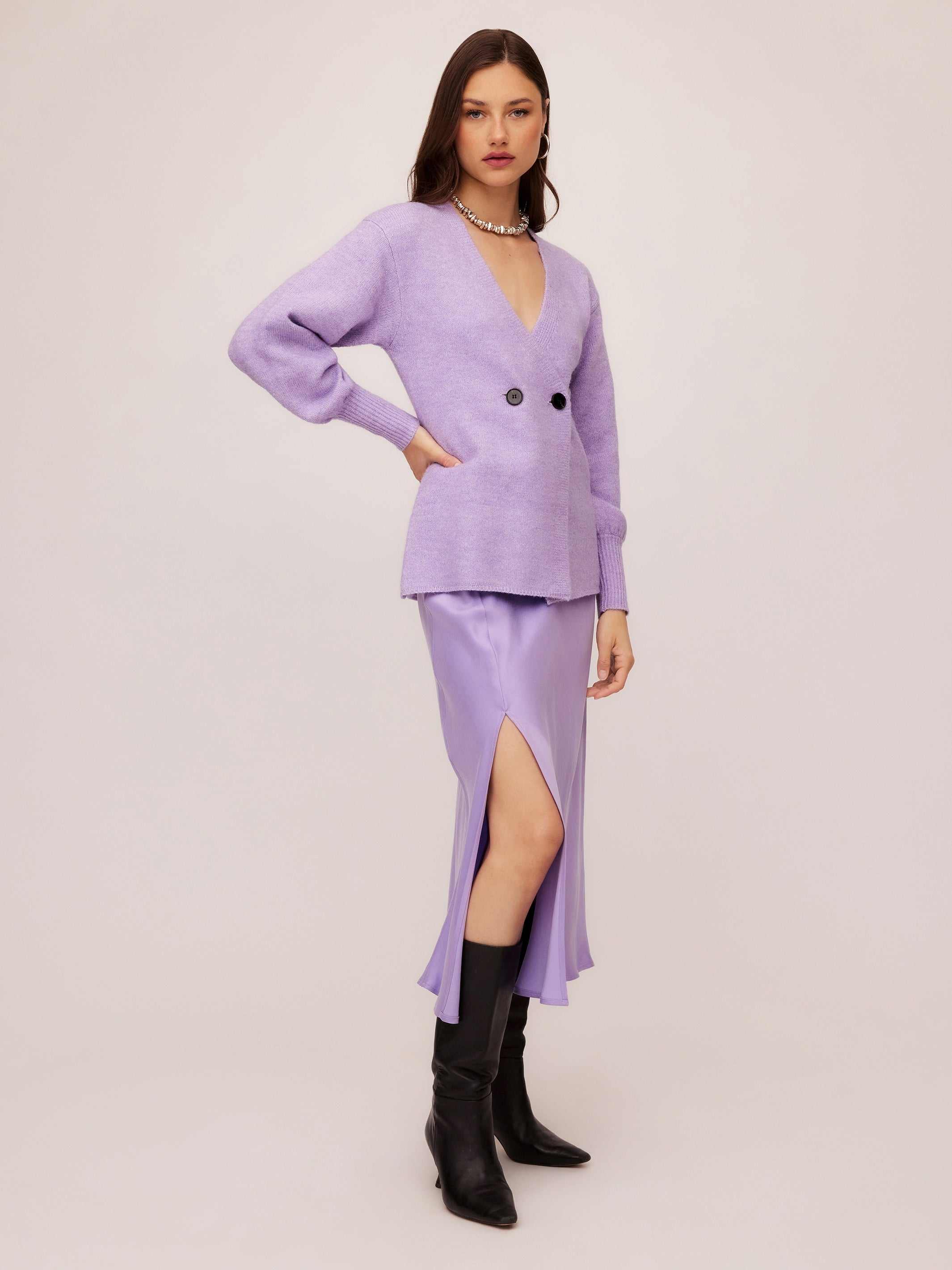 Woman wearing lavender Lucy Cardigan with double-breasted buttons, wide cuffs, and V-neck, paired with a lilac slit skirt and black boots