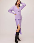Woman wearing a lilac Lucy Cardigan with double-breasted buttons, wide cuffs, and V-neck paired with a matching slit skirt and black boots