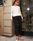 Woman wearing Luca Crewneck Sweater with beige stripes, black pants, and white heels standing by a decorative iron gate