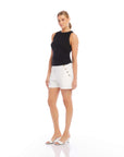 woman wearing black side shirred tank with white shorts and white sandals standing against white background