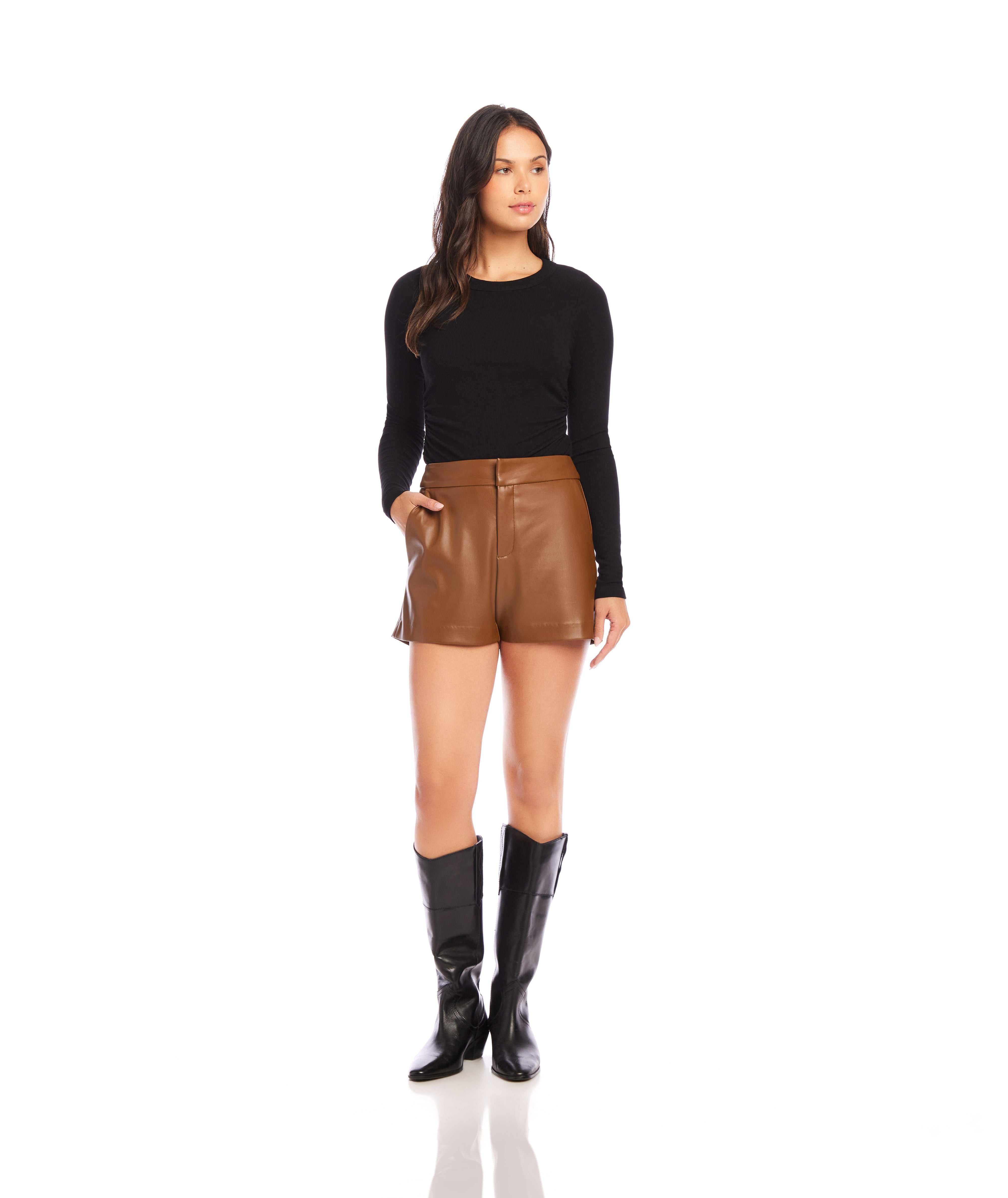 Woman wearing black long sleeve side shirred top with brown leather shorts and black boots standing on white background