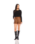 Woman wearing black long sleeve side shirred top with brown leather shorts and black boots standing on white background