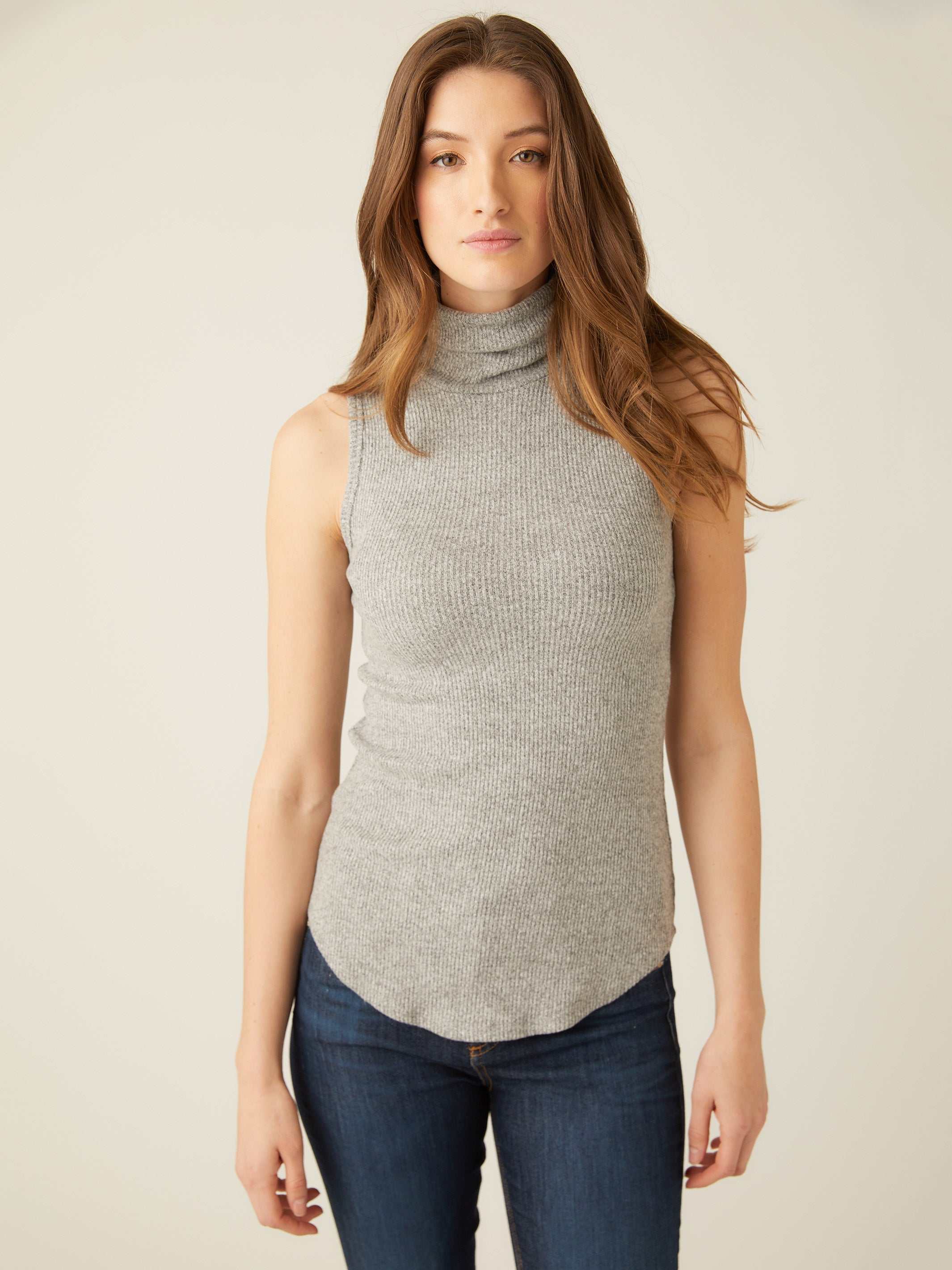 Woman wearing grey sleeveless turtleneck with rib-knit texture and shirttail hem paired with jeans