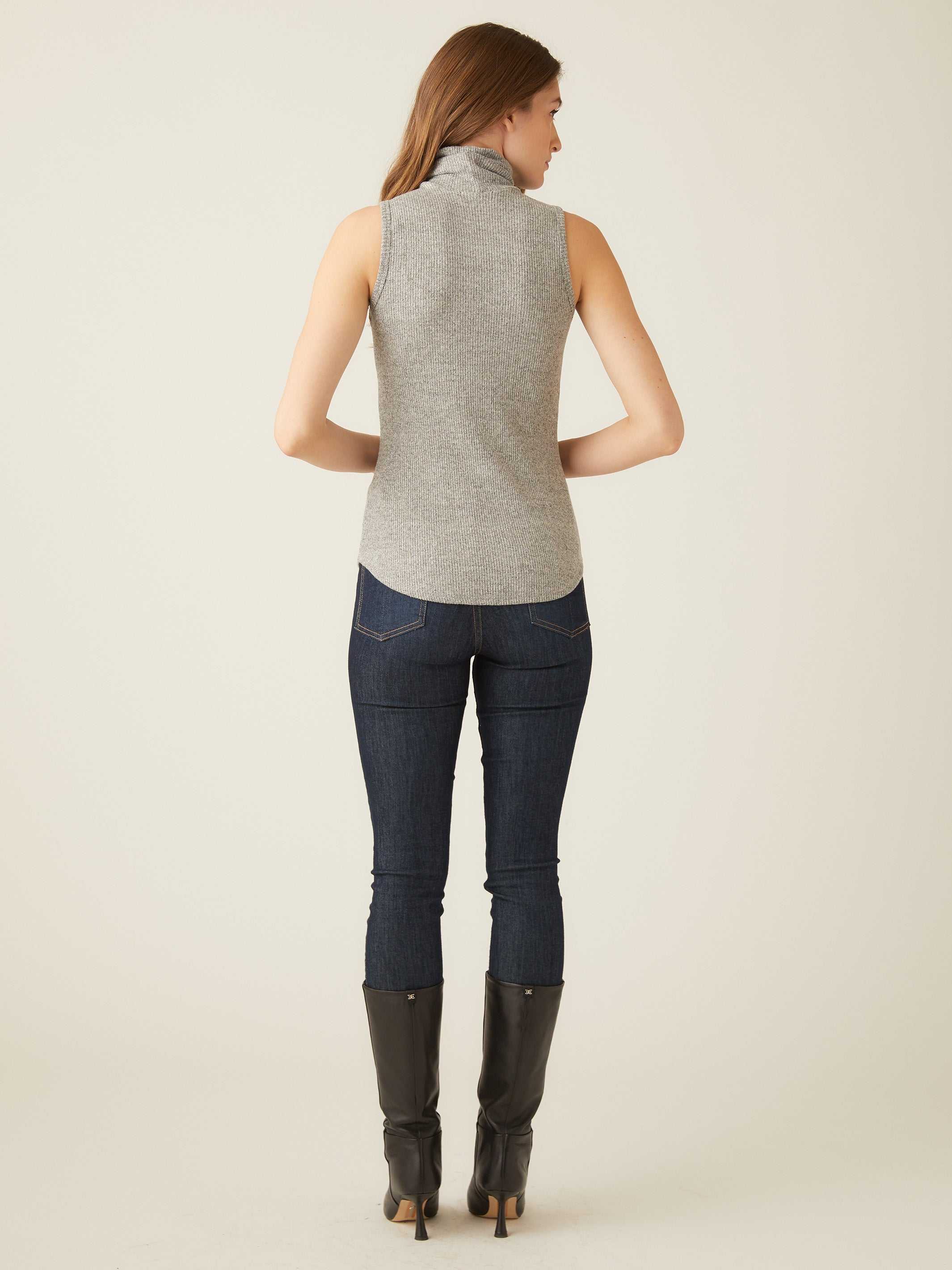 Woman wearing grey sleeveless turtleneck top with shirttail hem paired with dark jeans and black boots, back view
