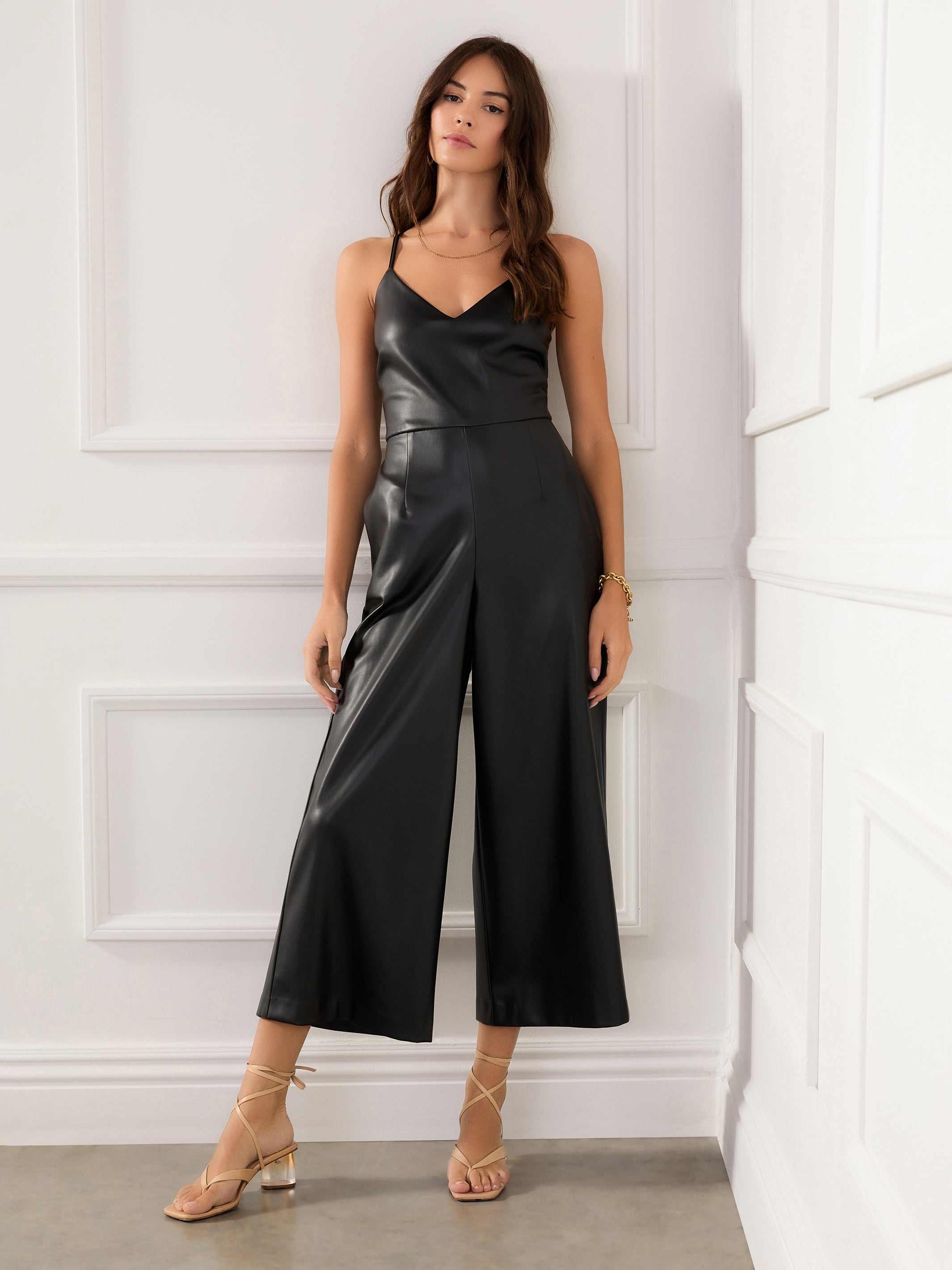 Woman wearing black fitted waist faux leather cropped jumpsuit with adjustable spaghetti straps and loose legs in a white room