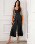 Woman wearing black fitted waist faux leather cropped jumpsuit with adjustable spaghetti straps and loose legs in a white room