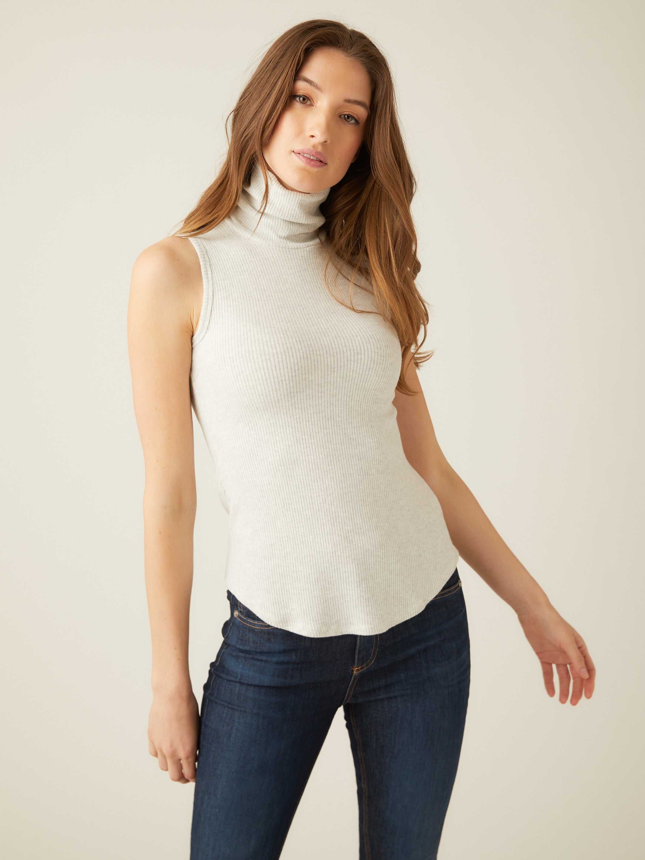 Woman wearing a slim fit sleeveless turtleneck rib-knit top with shirttail hem and dark jeans
