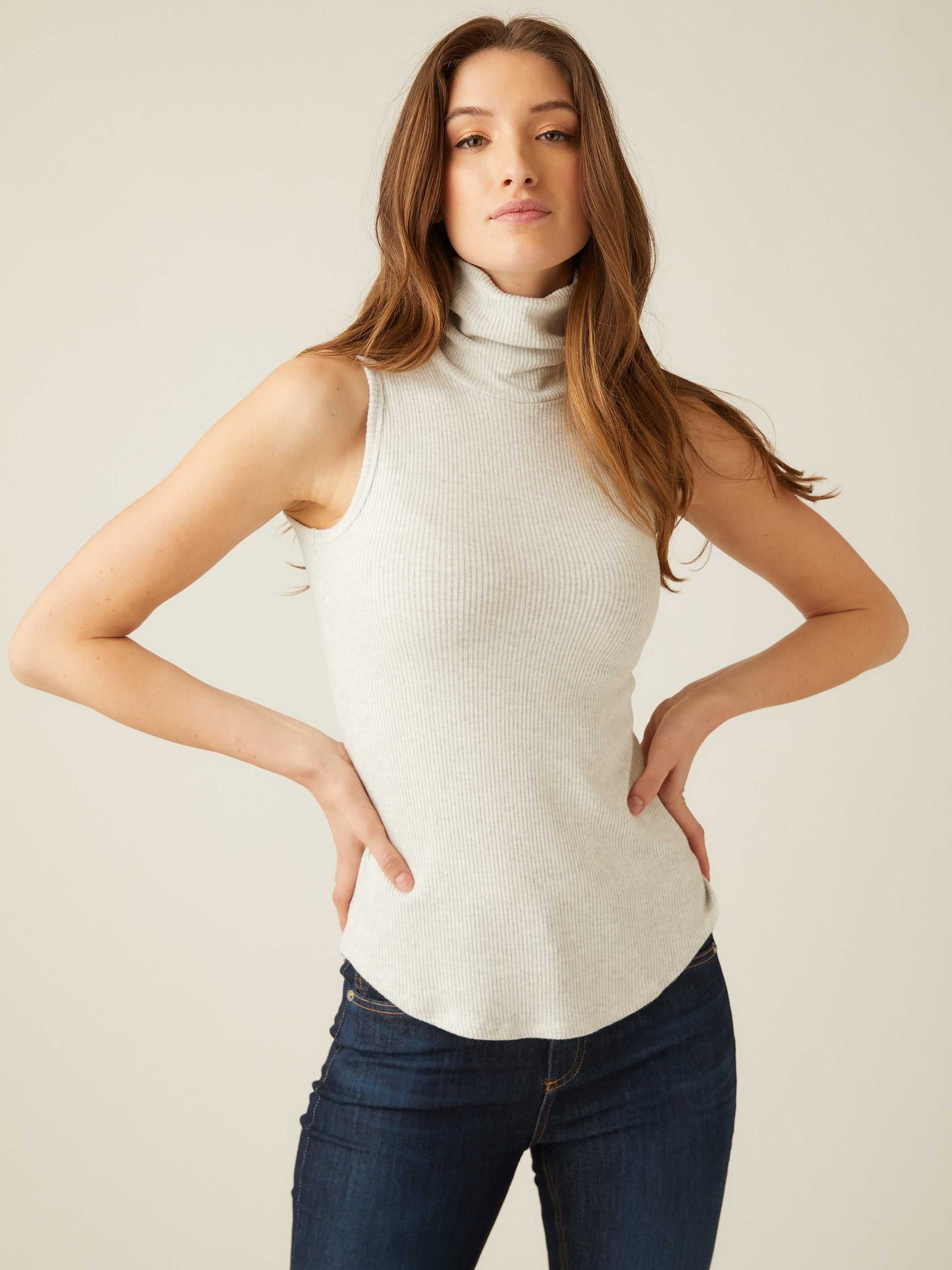 Woman wearing a slim fit sleeveless turtleneck rib-knit top in light gray with shirttail hem.