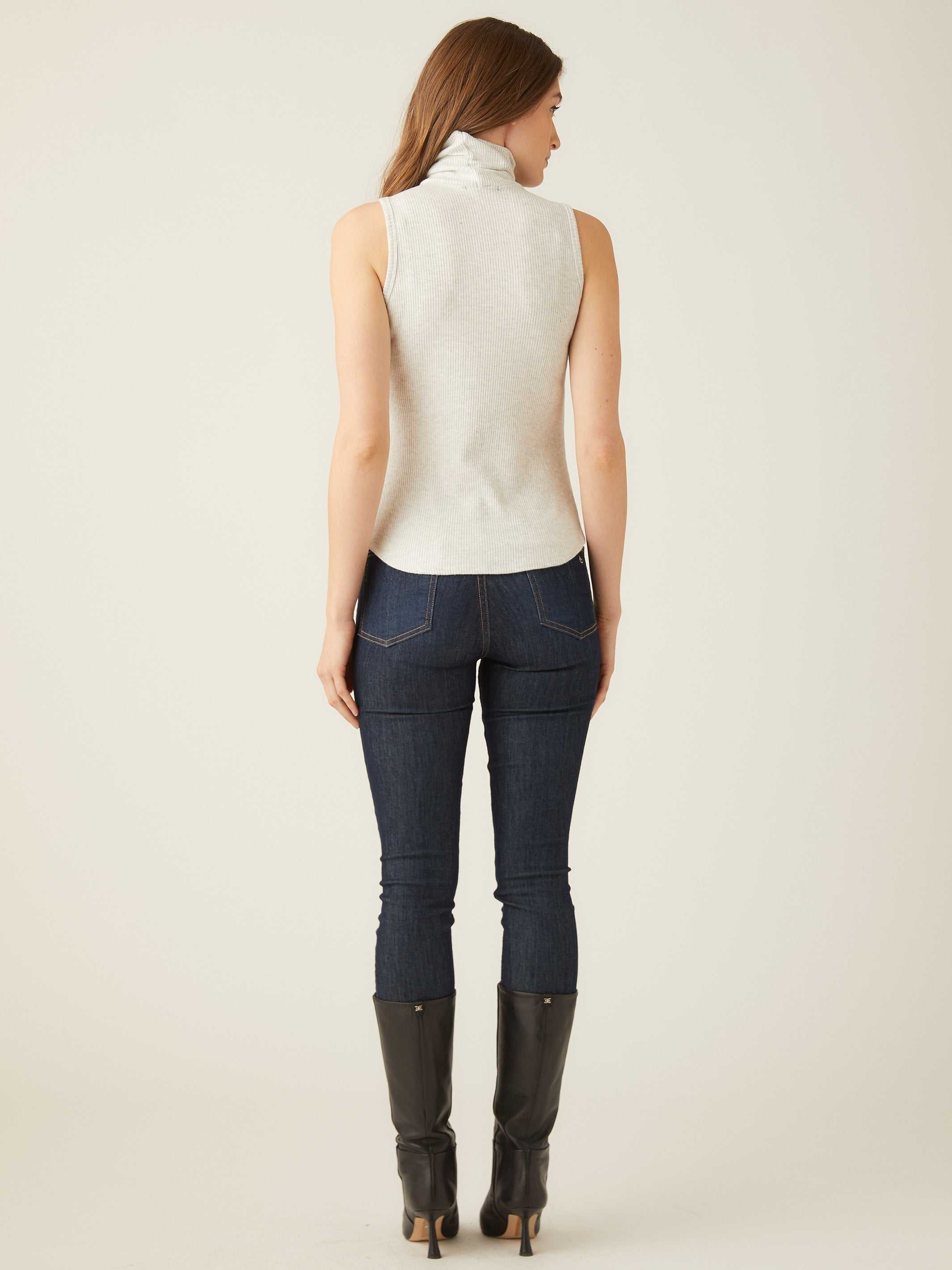 Woman wearing a slim fit sleeveless turtleneck top paired with dark jeans and black boots, back view