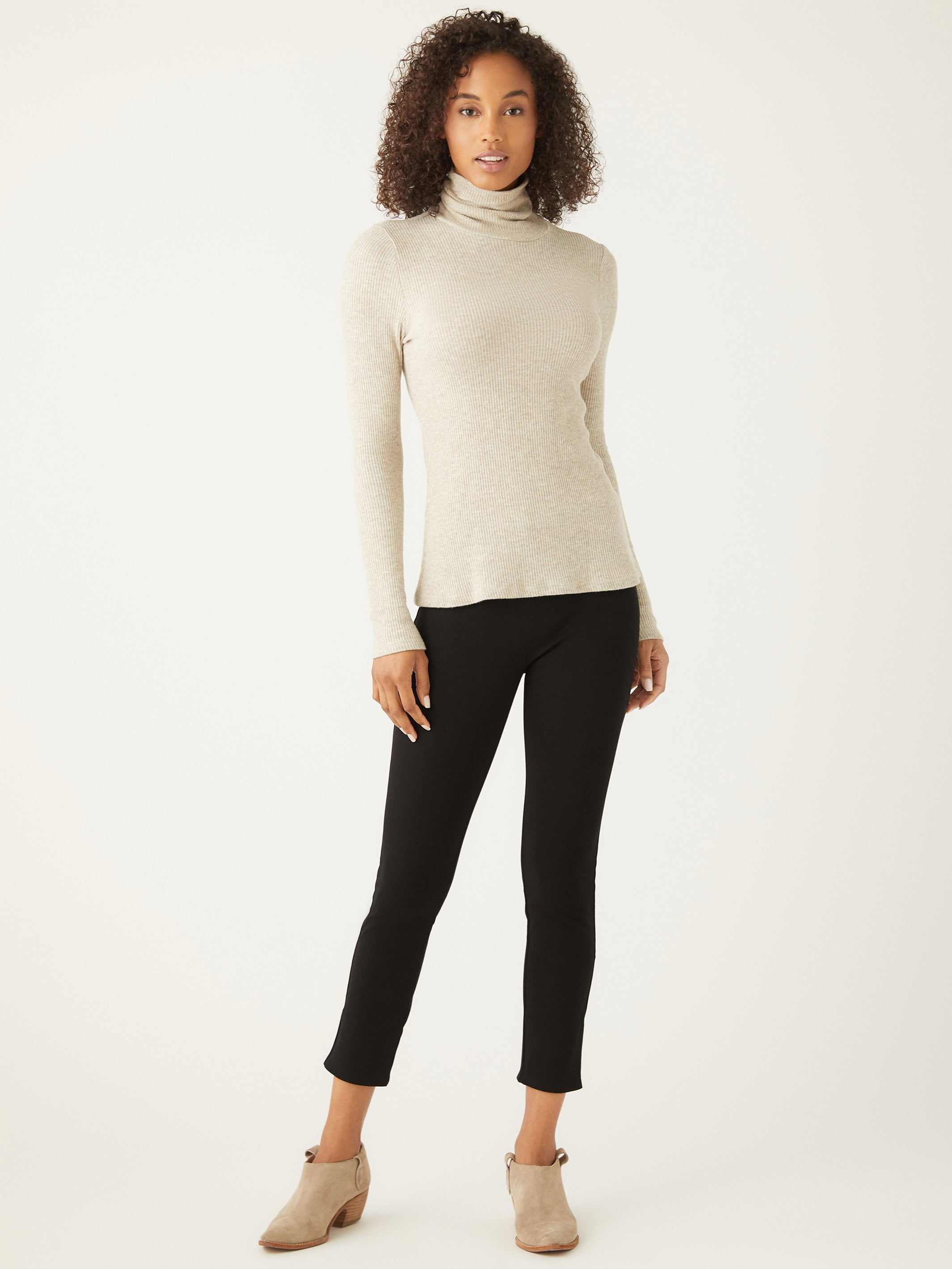 Woman wearing beige long sleeve turtleneck sweater and black pants standing against white background