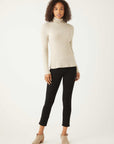 Woman wearing beige long sleeve turtleneck sweater and black pants standing against white background