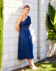 Woman wearing blue Abilene One-Shoulder Midi Dress with puff sleeve and back slit, standing against tiled wall outdoors