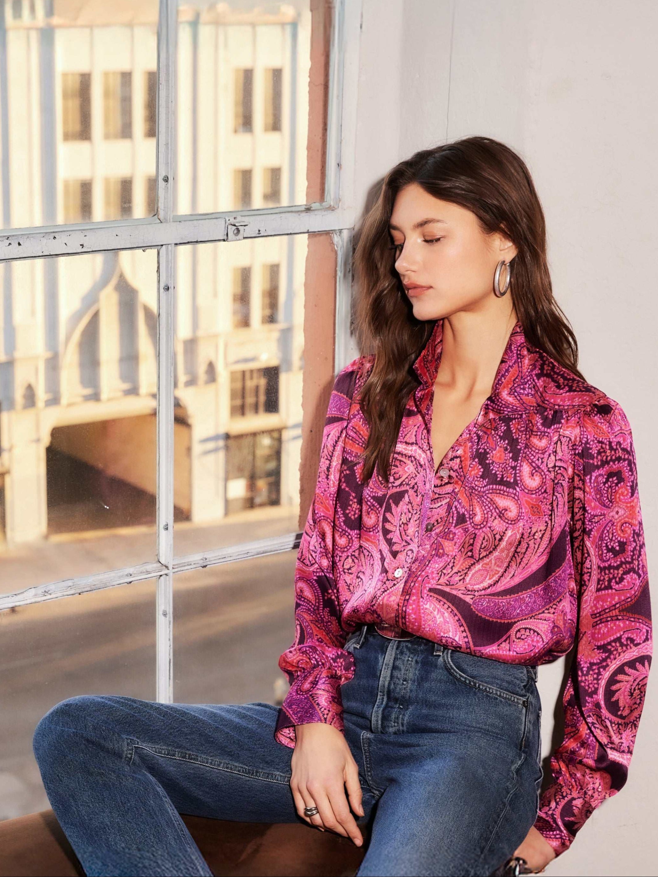 Woman wearing a pink paisley crinkle satin Chloe Blouse with gathered shoulders and buttoned cuffs paired with blue jeans sitting by a window.