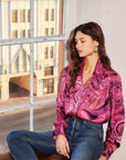 Woman wearing a pink paisley crinkle satin Chloe Blouse with gathered shoulders and buttoned cuffs paired with blue jeans sitting by a window.