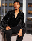 Woman wearing black Bianca Collared Shirred Top in crushed velvet with long sleeves and button cuffs sitting indoors by window
