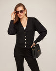 Woman wearing black Madeline Cardigan with V-neck, gathered shoulders, pearly buttons, and wide hem band.